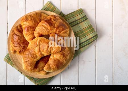 Frisches Croissant auf gelber und grüner karierter Serviette und Weidenplatte auf rustikalem Hintergrund aus weißem Holz. Overhead-Ansicht mit Platz für Text. Stockfoto