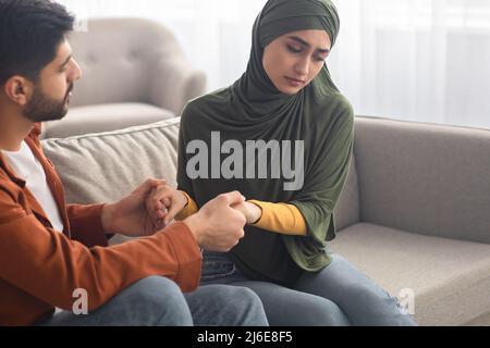 Unglückliches Paar Aus Dem Nahen Osten, Das Hände Auf Dem Sofa Im Wohnzimmer Hält Stockfoto