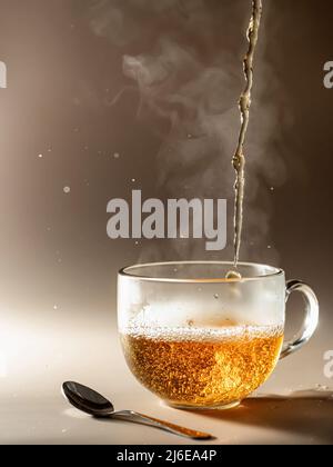 Tee wird in Glas-Teetasse mit Dampf und Spritzern auf braunem monochromen Hintergrund gegossen Stockfoto