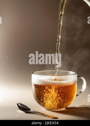 Tee wird in Glas-Teetasse mit Dampf auf braunem monochromen Hintergrund gegossen Stockfoto