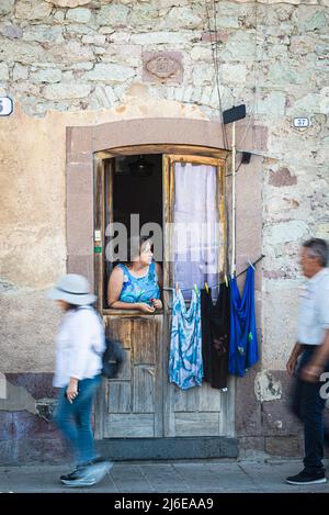 Sardisches Straßenleben - eine ältere Frau schaut aus der Eingangstür nach rechts auf die Straße, während zwei Fußgänger auf dem Bürgersteig vorbeikommen Stockfoto