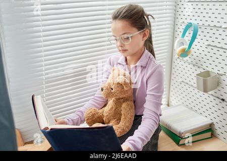 Konzentrierte kluge Mädchen in Gläsern sitzen mit Spielzeug Bär auf den Knien und Buch zu lesen, um es Stockfoto