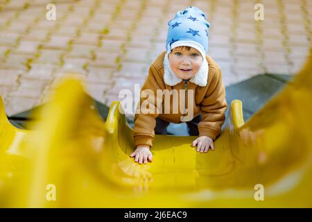 Fröhlicher lustiger kleiner Junge in einem blauen Hut und warmer Kleidung spielt auf dem Spielplatz und reitet von einer hellen bunten Rutsche in einem Kindergarten, in einem Stockfoto