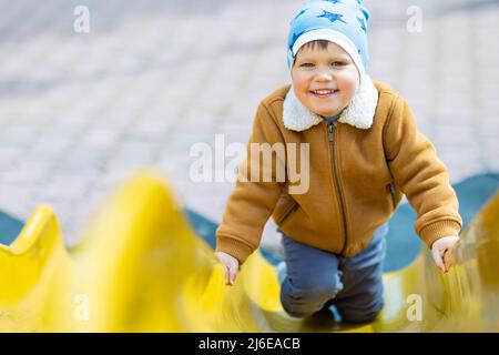 Fröhlicher lustiger kleiner Junge in einem blauen Hut und warmer Kleidung spielt auf dem Spielplatz und reitet von einer hellen bunten Rutsche in einem Kindergarten, in einem Stockfoto