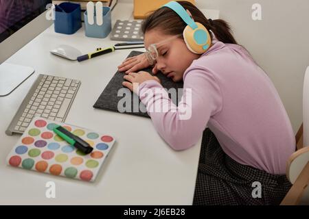 Müde Mädchen in kabellosen Kopfhörern und Brillen schlafen auf dem Schreibtisch mit Computer und Copybooks Stockfoto