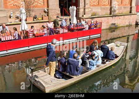 Colmar, Frankreich - 2022. April: Touristen in einem kleinen Boot auf dem Kanal an Menschen vorbeifahren, die an Tischen eines Cafés sitzen Stockfoto