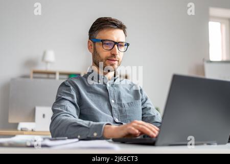 Fokussierter, reifer Geschäftsmann, der mit einem Laptop an seinem Schreibtisch im Büro arbeitet, auf der pc-Tastatur tippt und E-Mails checkt Stockfoto