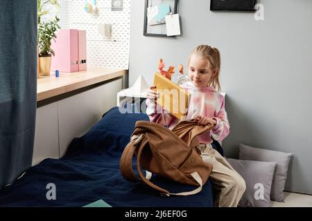 Nettes blondes Mädchen, das auf dem Bett sitzt und das Buch in die Tasche legt, während es sich für die Schule vorbereitet Stockfoto