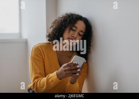 Unglückliche junge schwarze Frau, die schreckliche Nachrichten auf dem Handy liest und das Internet zu Hause übernutzt Stockfoto