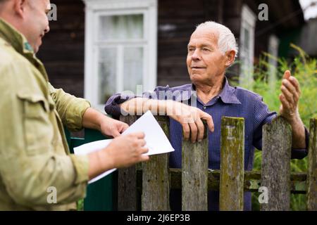 Leitender Mann, der mit dem Vertreter der Versicherungsgesellschaft kommuniziert, während er am Zaun seines Landhauses steht Stockfoto