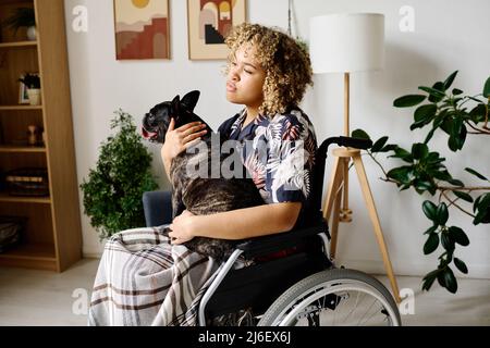 Afrikanisches junges Mädchen mit Behinderung sitzt auf dem Rollstuhl und umarmt den Hund, der auf den Knien im Zimmer sitzt Stockfoto