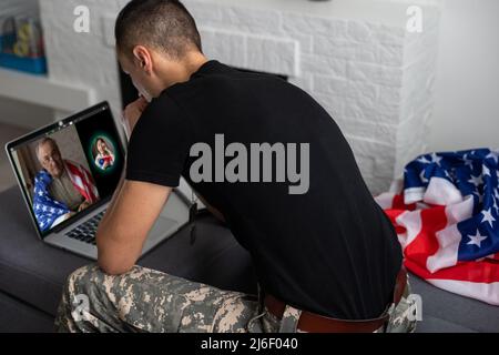 Patriotischer Militärangehöriger im Headset, der in der Nähe der amerikanischen Flagge auf einen Laptop schaut Stockfoto