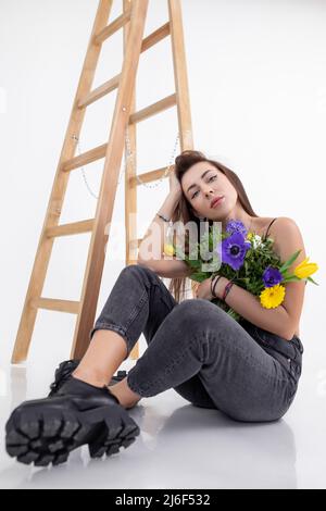 Porträt einer jungen hübschen Frau mit langen dunklen Haaren, die graue Jeans trägt, in der Nähe einer Holzleiter sitzt und einen Strauß hält. Stockfoto