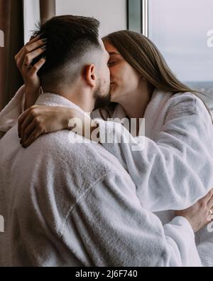 Glückliches, wunderschönes Paar im Hotel am Fenster küsst und umarmt sich gegenseitig Stockfoto