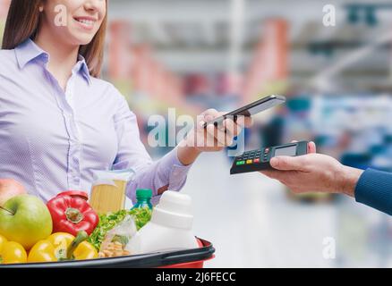 Frau, die mit ihrem Smartphone für Lebensmittel bezahlt, hält die Verkäuferin ein POS-Terminal Stockfoto