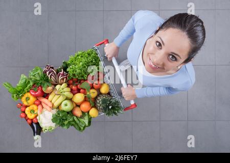 Glückliche junge Frau, die einen Trolley voller köstlichem Gemüse und Obst schiebt: Frische, gesunde Lebensmittel und Lebensmitteleinkaufskonzept Stockfoto