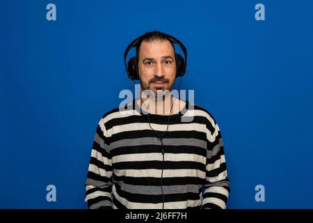 Hispanischer Mann mit einem Bart, der in einem gestreiften Pullover gekleidet ist und Musik mit Kopfhörern hört, isoliert auf blauem Studiohintergrund Stockfoto