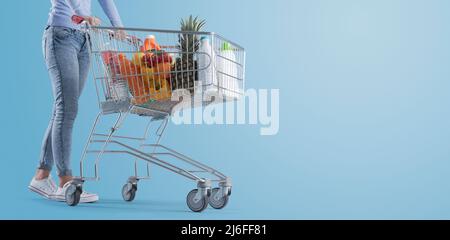 Junge Frau, die einen vollen Einkaufswagen, einen Supermarkt und ein Werbebanner für den Lebensmitteleinkauf schiebt, einen leeren Kopierplatz Stockfoto