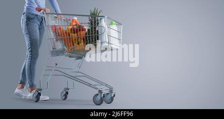 Junge Frau, die einen vollen Einkaufswagen, einen Supermarkt und ein Werbebanner für den Lebensmitteleinkauf schiebt, einen leeren Kopierplatz Stockfoto