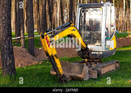 Ein kleiner Bagger gräbt einen Graben im Wald für Stromleitungen. Baumaschine Stockfoto