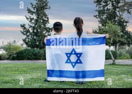 Kleine patriotische jüdische Kinder, die mit der Flagge Israels stehen und die Aussicht auf die Stadt genießen. Rückansicht Bild von jungen Kindern, die sich bei Sonnenuntergang mit der Flagge Israels auf der Wiese bedecken. Stockfoto