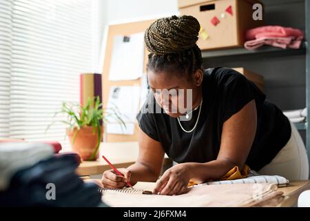 Junge ernsthafte schwarze Frau mit Bleistift über neue Skizze der Kleidung denken, während über die Schaffung der Sammlung von trendigen Kleidung arbeiten Stockfoto