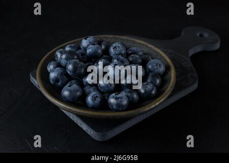 Frische Bio-Heidelbeeren in einer Keramikplatte. Die Platte steht auf einem grauen Brett auf schwarzem Hintergrund. Nahaufnahme. Stockfoto