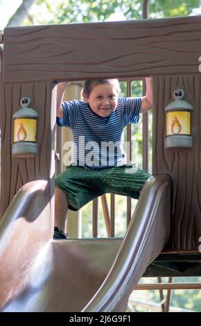 Der lachende Junge gucke aus dem oberen Teil einer Spielrutsche in einem Park heraus Stockfoto