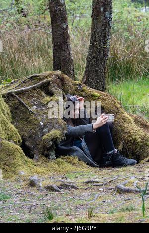 Junge Frau, die Selfie macht, während sie auf moosbedeckten Steinmauern in Schottland sitzt Stockfoto