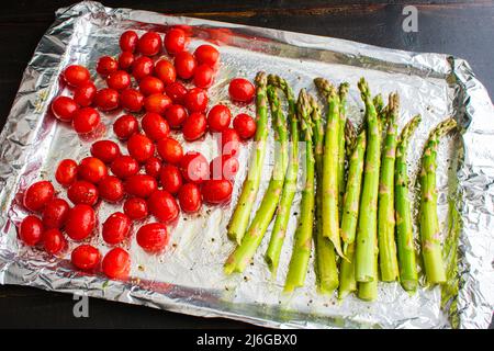 Frische Spargel- und Traubentomaten auf einer mit Folie überzogenen Blechpfanne: Eingestielter Spargel und ganze Traubentomaten, gewürzt mit Olivenöl, Salz und Pfeffer Stockfoto