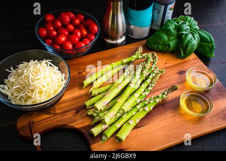 Eingestampfter Spargel auf einem Holzbrett mit anderen Zutaten: Spargel, Tomaten, Olivenöl und zerkleinerten Mozzarella-Käse Stockfoto