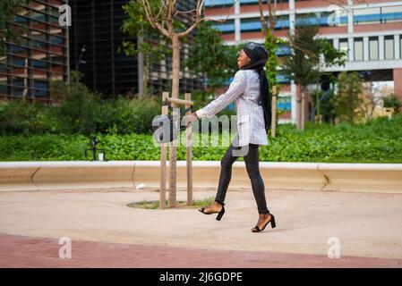 Fröhliche schwarze Frau im islamischen Kopftuch, die glücklich im Park läuft Stockfoto