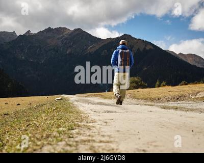 Rückansicht des asiatischen Wanderreisenden Rucksacktouristen, der auf einer unbefestigten Straße in Richtung Berge und Wald geht Stockfoto