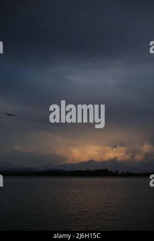 Ein Flugzeug klettert nach dem Start über das Meer, vor einer abgelegenen Gewitterwolke, die von der untergehenden Sonne beleuchtet wird Stockfoto