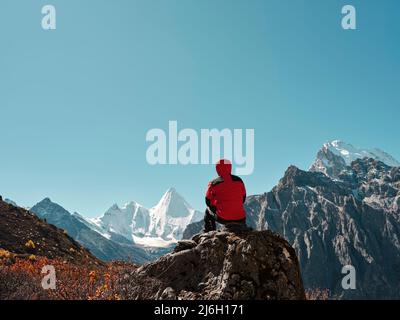 Rückansicht eines asiatischen Mannes, der auf einem Felsen mit dem Yangmaiyong (oder auf Tibetisch Jampayang)-Berggipfel in der Ferne in Yading, Landkreis Daocheng, sic sitzt Stockfoto