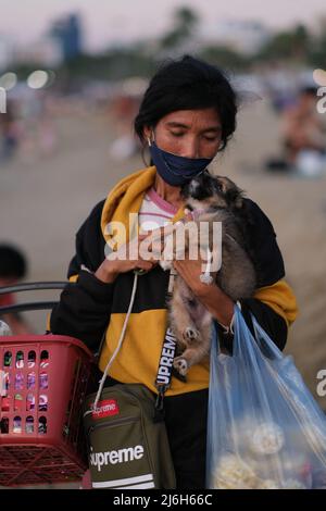 Pattaya, Chonburi Thailand - 11 2021. Dezember: Die arme Asiatin hält einen Welpen in der Hand, während sie am Strand Snacks verkauft Stockfoto