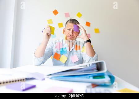 Der Mann sitzt hinter dem weißen Schreibtisch vor der Wand mit vielen bunten Notizen und hat auch viele Notizen auf sich und sucht nach einer Idee Stockfoto
