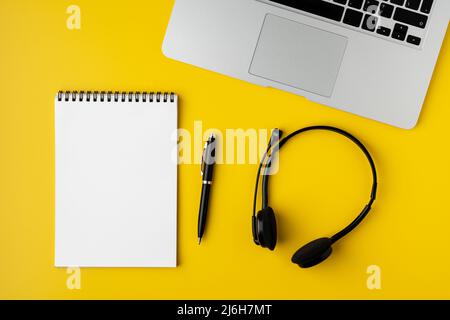 Moderner Büro-Desktop. Leerer Notizblock, Stift, Headset und Laptop auf gelbem Hintergrund Stockfoto