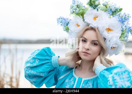 Schöne junge ukrainische Frau in einem blau bestickten Kleid mit einem großen schönen Blumenkranz auf dem Kopf gekleidet schaut auf die Kamera. Stockfoto