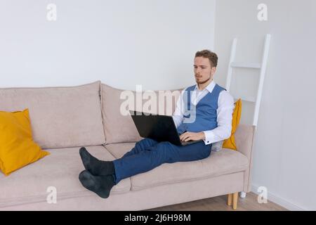 Portrait attraktiver, lächelnder junger bärtiger Mann in einem Business-Anzug sitzt im Wohnzimmer auf dem Sofa und arbeitet mit einem Laptop, der von zu Hause aus ferngesteuert arbeitet. Erfolgreicher kaukasischer Geschäftsmann Stockfoto
