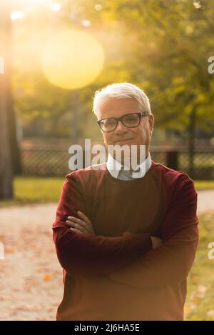 Vertikale Außenaufnahme eines selbstbewussten grauhaarigen kaukasischen Rentners im roten Pullover, der mit gekreuzten Armen im Park steht. Hochwertige Fotos Stockfoto