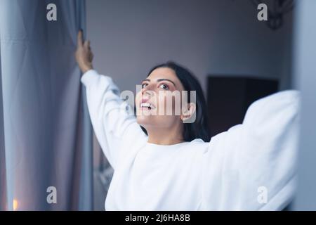 Fröhliche kaukasische schwarzhaarige Frau mit 30s in einem weißen Sweatshirt, die Fenstervorhänge öffnet und den neuen Tag begrüßt. Hochwertige Fotos Stockfoto