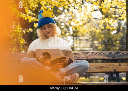 Schönes junges blondes kaukasisches Mädchen in einem Strickhut mit einem gelben Blatt, das ein Buch oder die Bibel auf einer Bank im Herbstpark liest. . Hochwertige Fotos Stockfoto