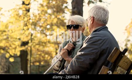 Ältere pensionierte kaukasische Ehepaar sitzt auf der Bank im Park, behinderte Frau in Brille und ein Stick Park selektive Fokus. Hochwertige Fotos Stockfoto