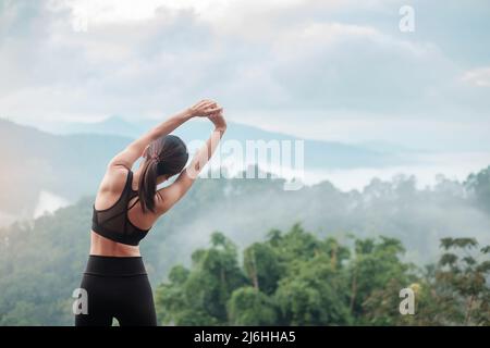 Junge Fitness-Frau in Sportbekleidung, die den Körper gegen den Blick auf die Berge streckt, gesunde Frau trainieren morgens. Training, Wohlbefinden und Work-Life-Balance Stockfoto