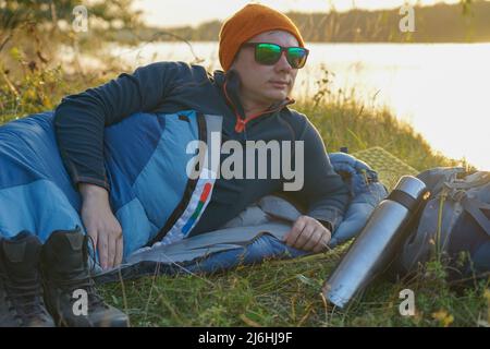 Guy Tourist liegt in einem Schlafsack am Ufer des Flusses. In der Nähe sind seine Sachen: Ein Rucksack, Stiefel, eine Thermoskanne, ein Teppich. Stockfoto