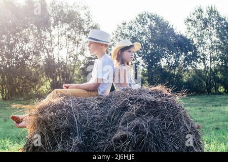 Porträt von zwei barfuß-Kindern, Jungen und Mädchen, die auf dem Heuhaufen auf dem Feld den Rücken zueinander sitzen. Kinder tragen Strohhüte. Leichter, sonniger Tag. Seite Stockfoto