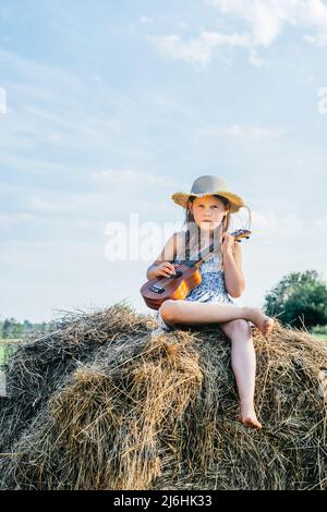 Porträt eines kleinen Mädchens, das kleine Ukulele spielt, auf dem Heuhaufen im Feld sitzt. Leichter, sonniger, wolkiger Tag. Fröhlich und genießen Konzept. Bäume an Stockfoto