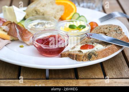 Frühstück mit Brot, Butter und Marmelade, auch verschiedene Aufschnitte und Früchte auf einem weißen Teller auf einem hölzernen Außentisch, ausgewählter Fokus, enge Fi-Tiefe Stockfoto