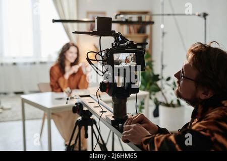 Junger Kameramann, der den Fokus und andere Optionen der Videokamera justiert, bevor er einen Werbefilm mit einer Frau am Tisch dreht Stockfoto